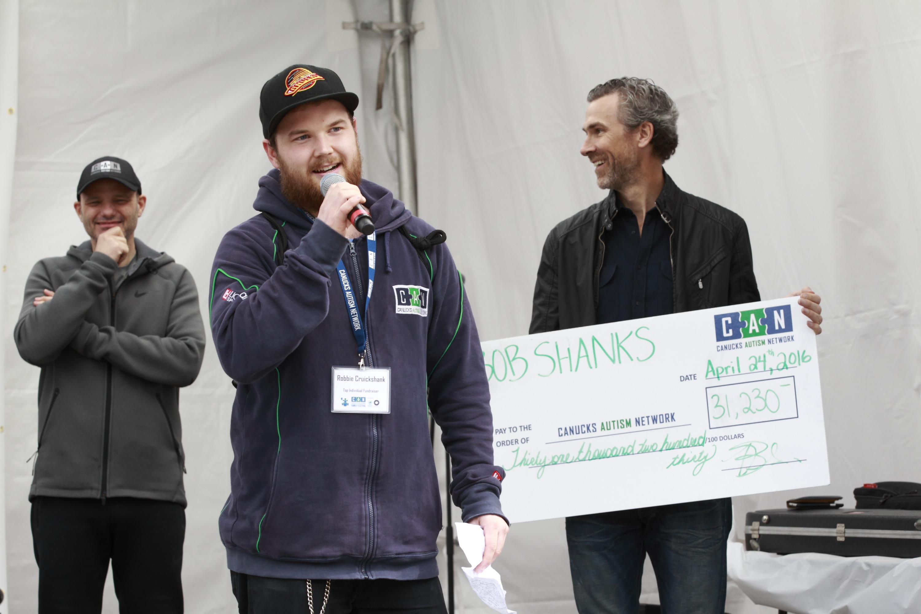 A young man speaks with a microphone with two men standing behind him holding a giant cheque.