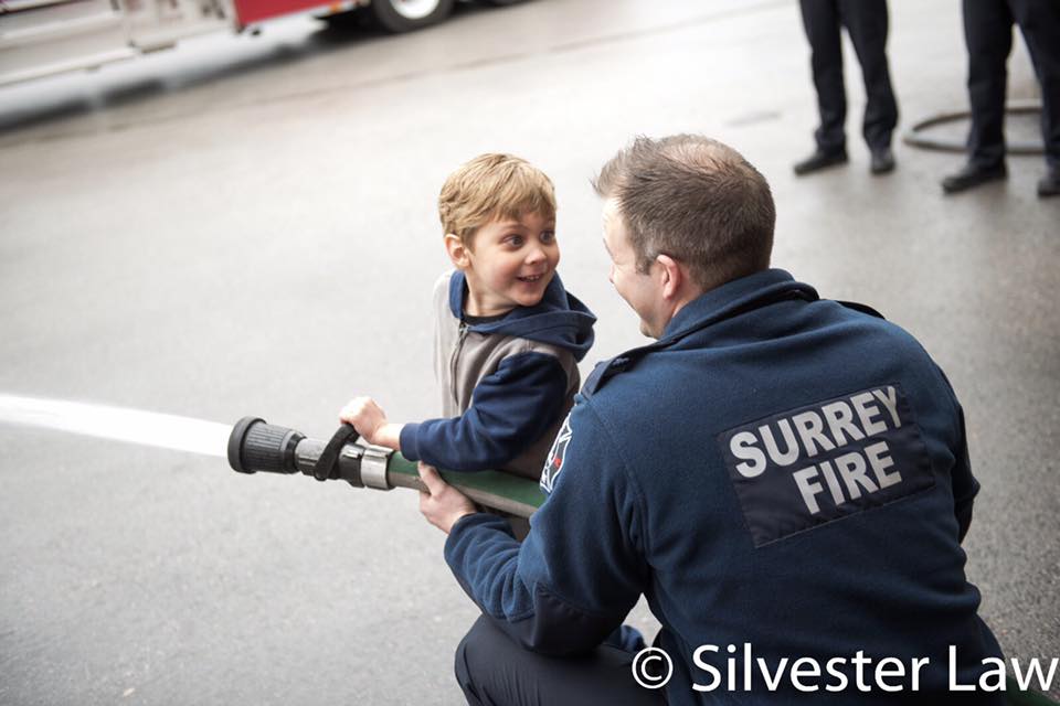 A child and a fire fighters hold a fire hose together, while the child looks back at the firefighter and smiles.