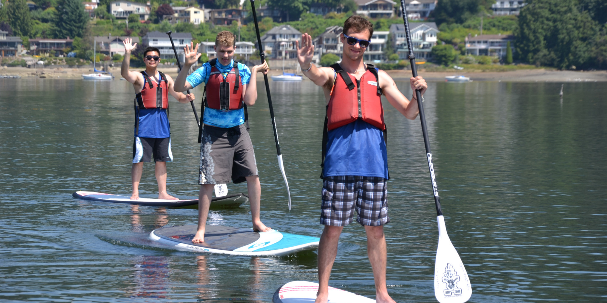 Three young men wave to the camera while standing on paddle boards.