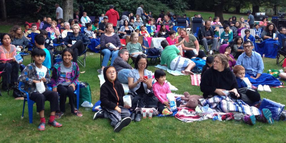 A group of roughly 100 families sit on blankets in the park.